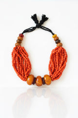 A handmade traditional coral and amber beaded necklace featuring two large, textured red-orange bead clusters of coral on either side, accented by three smooth orange-brown amber beads at the center, and smaller brown, yellow, and red beads at the top, all tied with a black cord on a neutral gray background.