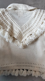 Close-up of a white knitted garment with a decorative neckline and tassels on a gray background.